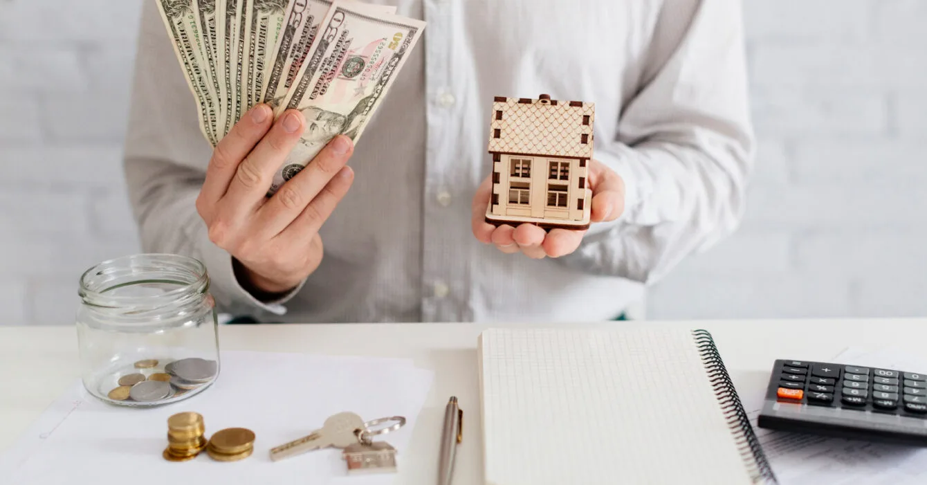 Real Estate Financing Thailand: Person holding money in one hand and a miniature house in the other over a desk with financial items.