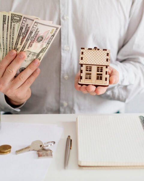 Real Estate Financing Thailand: Person holding money in one hand and a miniature house in the other over a desk with financial items.