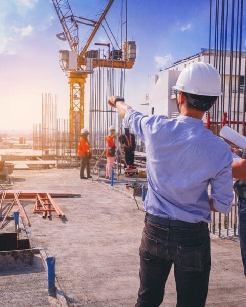 Two construction workers with helmets discussing plans on a building site with a crane in the background, symbolising new Construction Safety Thailand regulations.