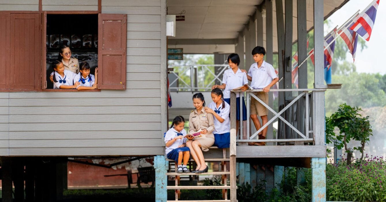 Thailand Urbanization Impact: Students in uniform at a wooden schoolhouse with Thai flags.