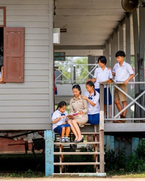 Thailand Urbanization Impact: Students in uniform at a wooden schoolhouse with Thai flags.